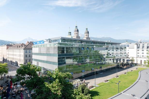 Das moderne Hauptgebäude des MCI Innsbruck mit Blick auf die umliegenden Berge und die Türme der Altstadt im Hintergrund.