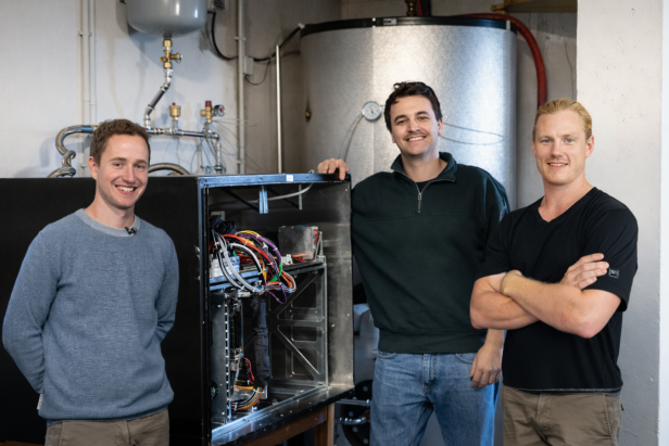 Drei Männer stehen vor einem offenen Serverschrank mit Kabeln und einem Warmwasserspeicher im Hintergrund.