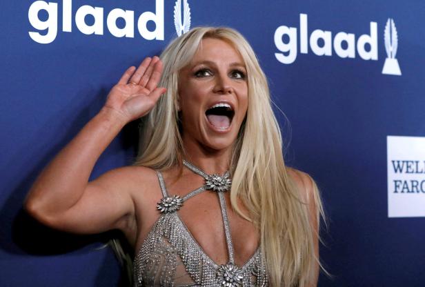 Britney Spears mit erhobener Hand und offenem Mund vor einem GLAAD-Hintergrund.