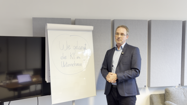 Mann im Anzug steht neben Flipchart mit der Aufschrift „Wie gelangt die KI ins Unternehmen?“.