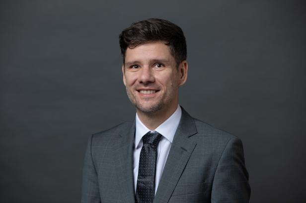 Ein Mann im Anzug steht lächelnd vor einer grauen Wand.