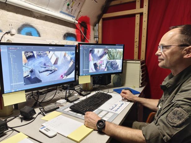 Ein Mann in Uniform überwacht mehrere Überwachungskameras an zwei Monitoren in einem Raum mit rotem Vorhang.