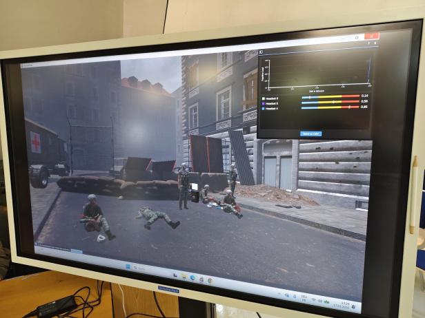 Mehrere Soldaten in Uniform befinden sich in einer Straßenszene mit Sandsäcken, Verletzten und einem Sanitätsfahrzeug im Hintergrund.