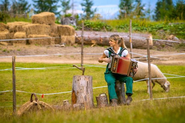 Eine Frau in grüner Latzhose spielt Akkordeon auf einer Wiese, während ein Schaf im Hintergrund grast.