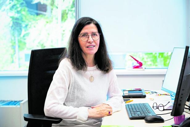 Eine Frau mit langen dunklen Haaren und Brille sitzt an einem Schreibtisch mit Computer und Tastatur vor einem Fenster.