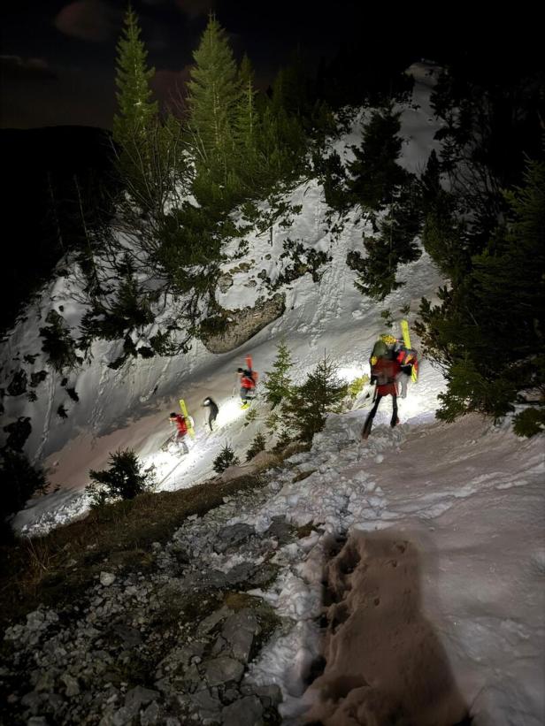 Vier Personen mit Stirnlampen und Ausrüstung steigen nachts einen verschneiten Hang im Gebirge hinauf.