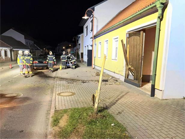 Mehrere Feuerwehrleute stehen auf einer nächtlichen Straße neben einem beschädigten Auto und einem zerstörten Holztor an einem Haus.