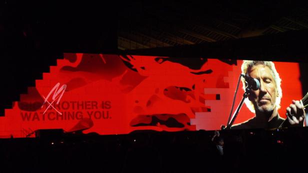 Roger Waters tritt vor einer roten Leinwand mit der Aufschrift „Big Brother is watching you“ auf.