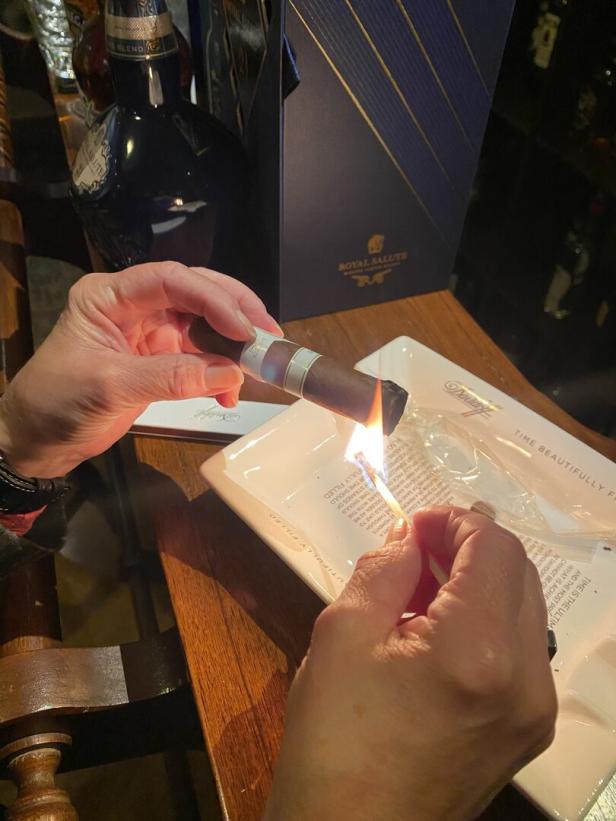Eine Person zündet eine Zigarre mit einem Streichholz an, daneben stehen ein Aschenbecher und eine Flasche Whisky.
