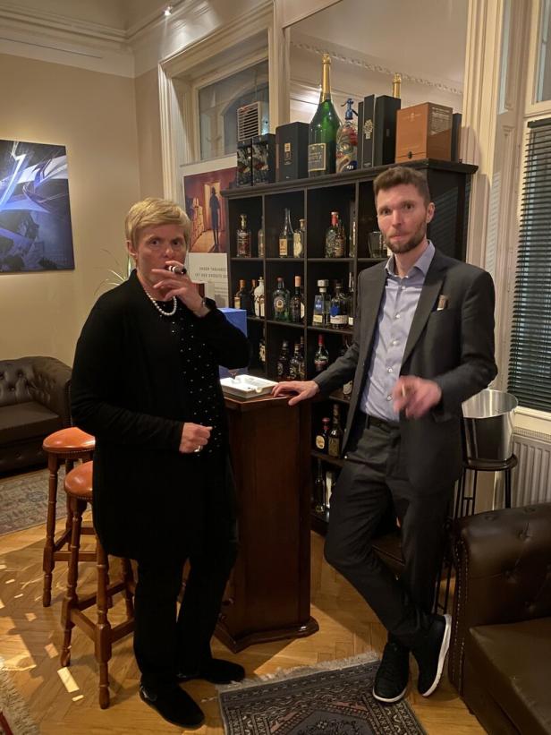 Zwei Personen stehen in eleganter Umgebung vor einem Regal mit Spirituosen und rauchen Zigarren.