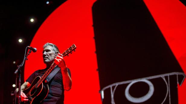 Roger Waters spielt Gitarre auf einer Bühne mit roter Beleuchtung.