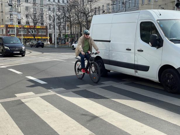 Ein Mann mit Helm fährt auf einem Fahrrad neben einem weißen Lieferwagen über einen Zebrastreifen in der Stadt.