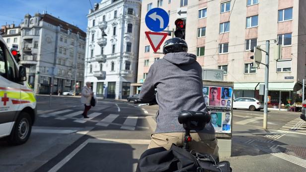 Ein Radfahrer mit Helm wartet an einer roten Ampel an einer Kreuzung, links steht ein Rettungswagen, im Hintergrund sind Gebäude.