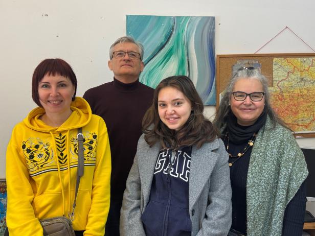 Vier Personen stehen nebeneinander und lächeln, im Hintergrund sind eine Landkarte und ein abstraktes Gemälde zu sehen.