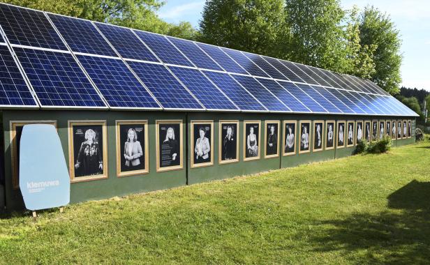Eine lange grüne Wand mit vielen gerahmten Schwarzweißporträts und darüber große Solarpaneele im Freien.