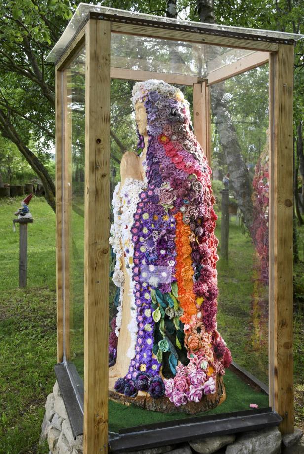 Holzskulptur einer stehenden Figur, mit bunten gehäkelten Blumen bedeckt, steht in einem gläsernen Schutzkasten im Grünen.