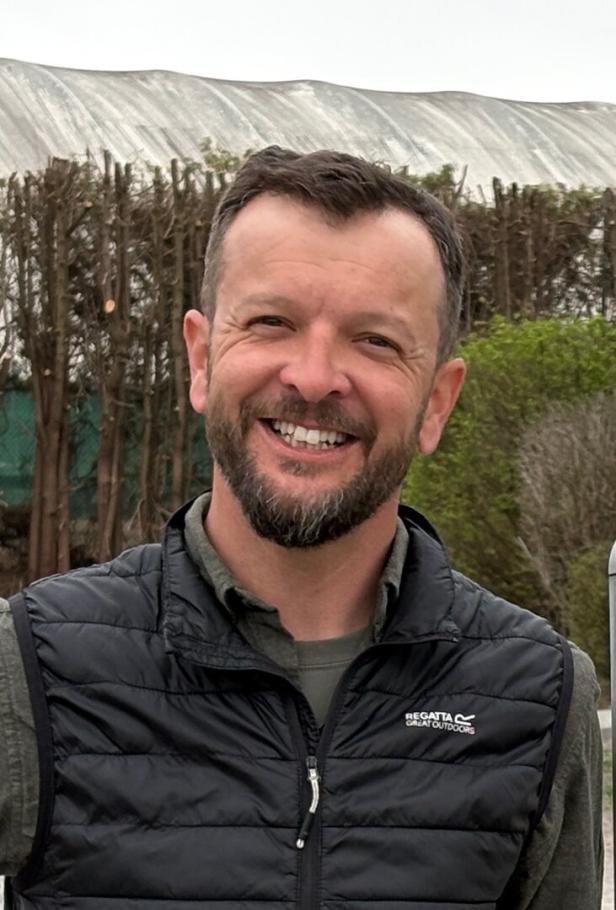 Ein Mann mit Bart und kurzer Frisur lächelt im Freien, trägt eine schwarze Steppweste und ein grünes Hemd.