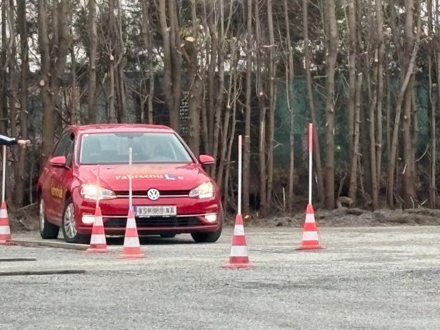 Ein rotes Auto mit Fahrschulaufschrift fährt zwischen Verkehrshütchen auf einem Übungsplatz vor kahlen Bäumen.