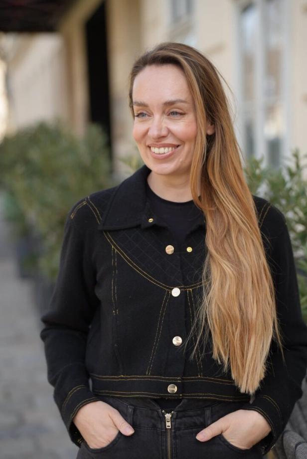 Frau mit langen blonden Haaren und schwarzer Jacke steht draußen und lächelt, Hände in den Hosentaschen.