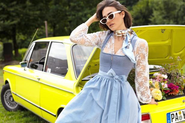 Frau in blauem Dirnd lehnt an einem gelben Auto