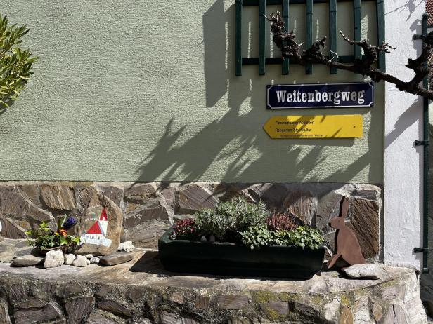 Straßenschild „Weitenbergweg“, gelber Wegweiser, Blumenkasten, bemalter Stein und Hasenfigur an einer Mauer.