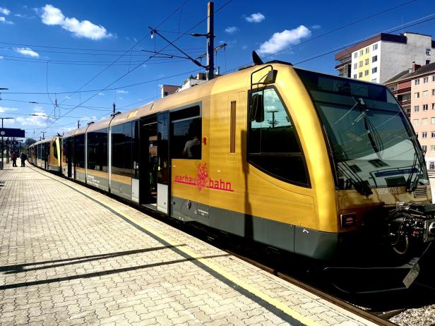 Ein goldfarbener Zug mit der Aufschrift „wachau bahn“ steht an einem sonnigen Bahnhof neben mehrstöckigen Wohnhäusern.