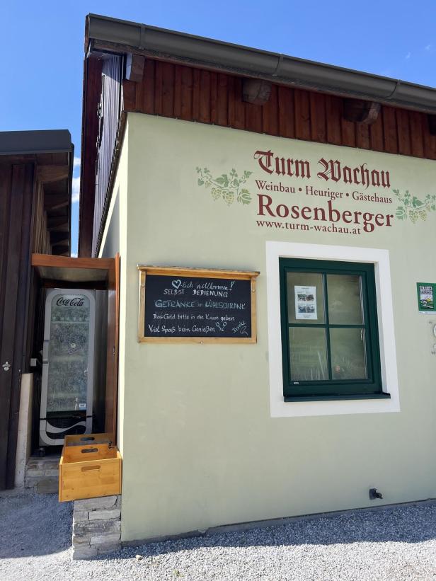 Hausfassade mit grüner Tür und Fenster, daneben eine Tafel mit Infos zur Selbstbedienung und ein Getränkekühlschrank im Eingangsbereich.