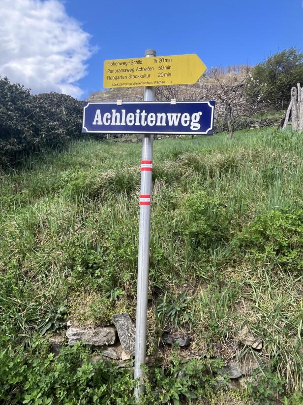 Wegweiser mit der Aufschrift "Achleitenweg" vor einer grünen Wiese und blauem Himmel mit Wolken.