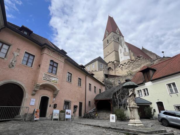 Historische Gebäude mit einer Statue im Vordergrund und einer Kirche mit rotem Dach im Hintergrund unter bewölktem Himmel.