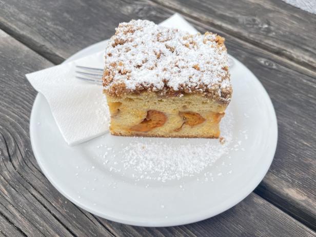 Ein Stück Streuselkuchen mit Aprikosen und Puderzucker auf einem weißen Teller mit Gabel und Serviette.