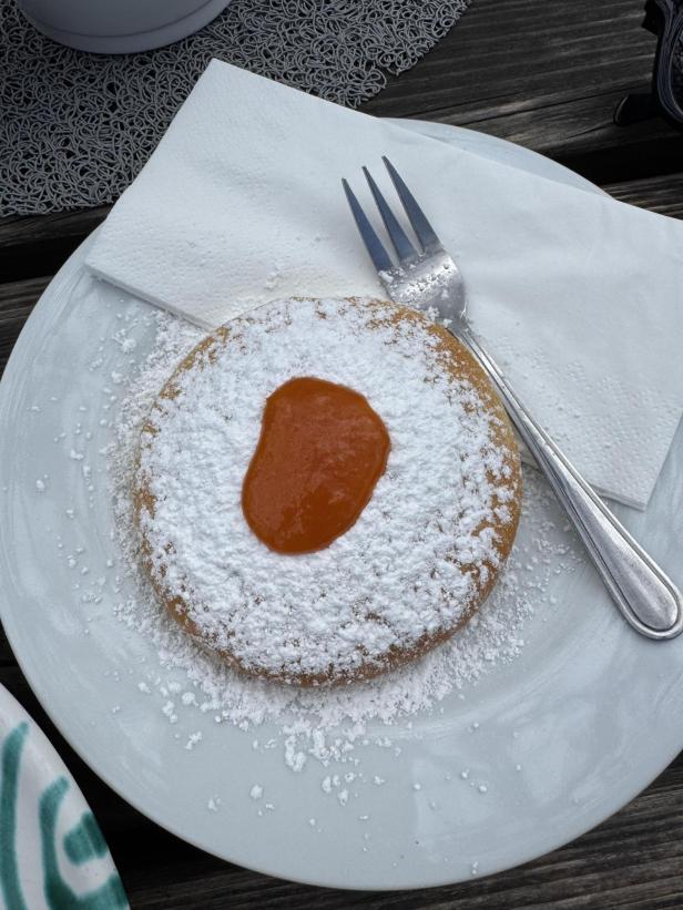 Ein rundes Gebäck mit Puderzucker bestreut und einem Klecks Marmelade in der Mitte liegt auf einem weißen Teller mit Gabel und Serviette.