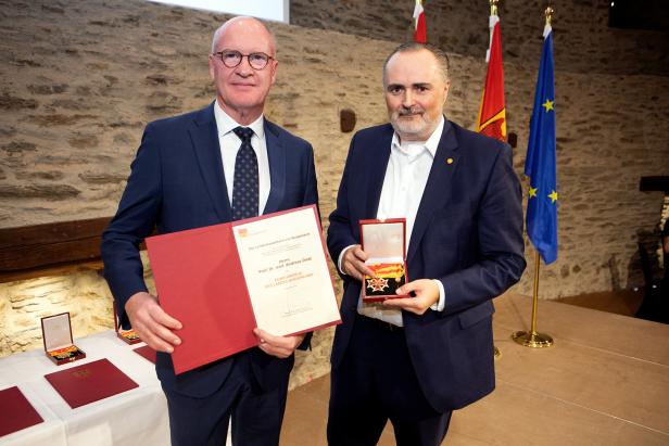 Zwei Männer im Anzug halten eine Urkunde und eine Auszeichnung, im Hintergrund stehen Flaggen und ein Steintisch.