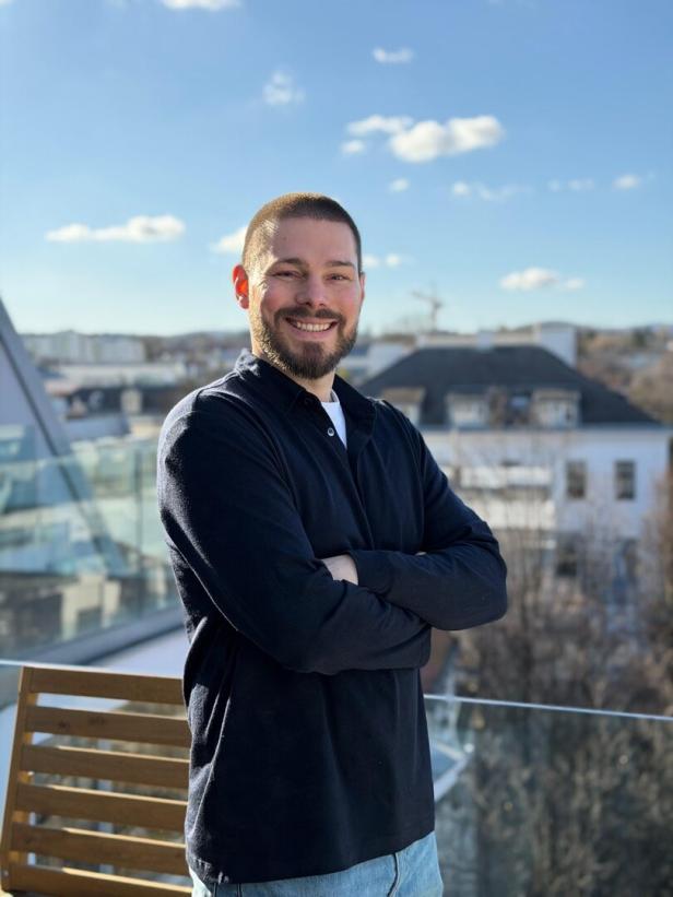 Ein Mann steht lächelnd auf einer Terrasse mit Häusern im Hintergrund.