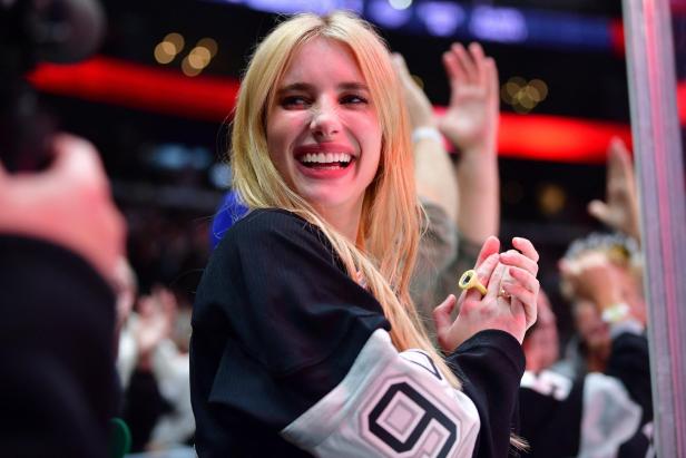 Emma Roberts mit blondem Haar lacht und klatscht inmitten einer jubelnden Menschenmenge bei einer Sportveranstaltung.