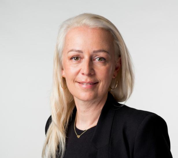 Eine Frau mit blonden Haaren, goldenen Ohrringen und einer schwarzen Jacke lächelt.