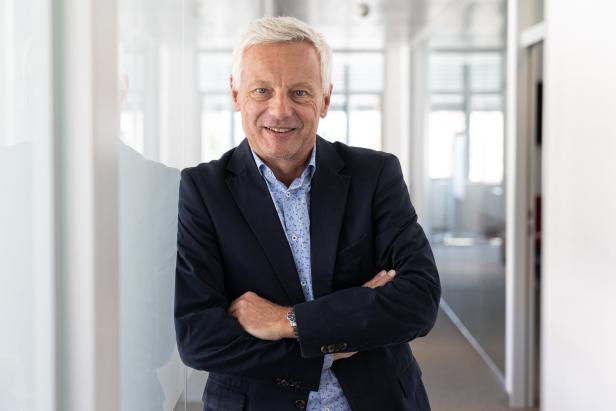 Ein älterer Mann mit grauen Haaren steht mit verschränkten Armen in einem modernen, hellen Flur und lächelt freundlich.