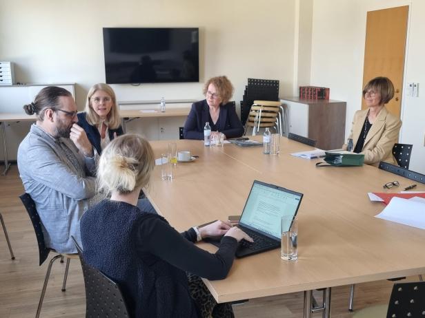 Vier Personen sitzen um einen Tisch und unterhalten sich, vor ihnen Gläser, Kaffeetassen und ein Laptop.