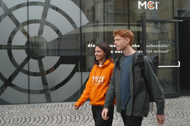 Zwei Studierende gehen lachend am Eingang des MCI Innsbruck vorbei, das moderne Hochschulgebäude und das MCI-Logo sind im Hintergrund sichtbar.