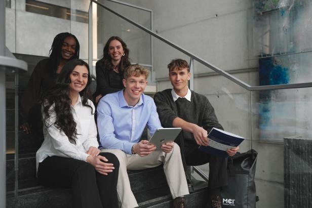 Fünf internationale Studierende des MCI Innsbruck sitzen gemeinsam auf einer Treppe und lächeln, mit Lernmaterialien und Tablet in der Hand.