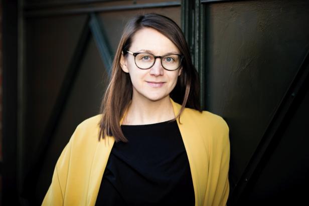 Frau mit braunen Haaren und Brille trägt einen gelben Blazer über einem schwarzen Oberteil und blickt freundlich nach vorn.