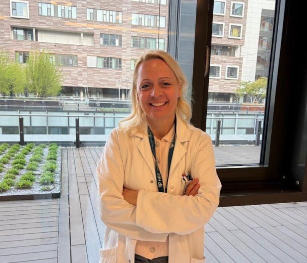 Eine Frau im weißen Kittel steht lächelnd mit verschränkten Armen vor einem Fenster mit Blick auf moderne Gebäude und eine Terrasse.