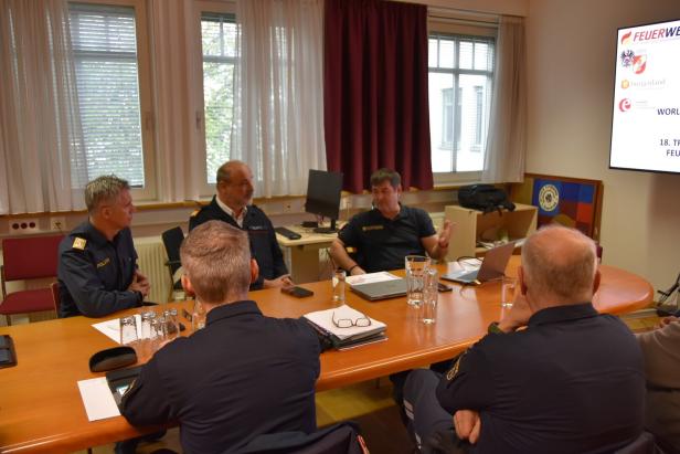 Fünf Männer in Uniformen sitzen an einem Konferenztisch und besprechen sich, auf einem Bildschirm steht „FEUERWEHR“.