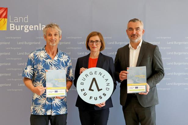 Drei Personen stehen vor einer Wand mit „Land Burgenland“-Schriftzug und halten Broschüren sowie ein rundes Schild mit „Burgenland zu Fuß“.