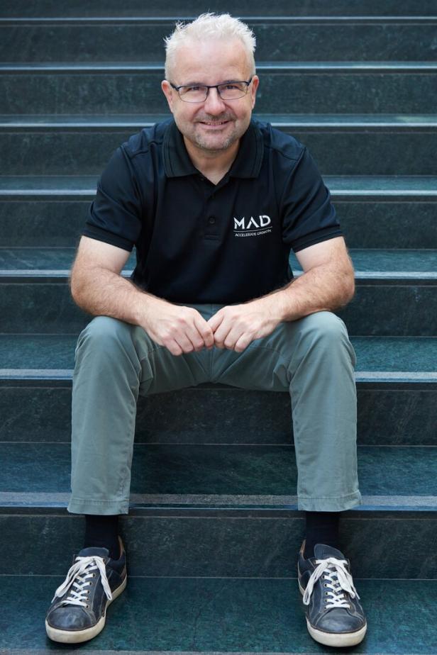 Ein Mann mit Brille und grauen Haaren sitzt lächelnd auf einer Treppe und trägt ein schwarzes Poloshirt sowie Turnschuhe.