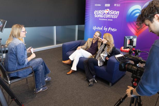 Drei Personen führen ein Interview vor einer Eurovision Song Contest 2026 Wand, während ein Kameramann die Szene filmt.