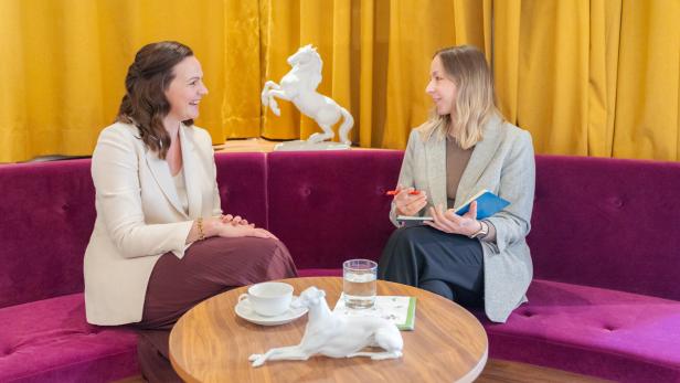 Zwei Frauen sitzen auf einem lila Sofa und unterhalten sich an einem Tisch mit Getränken und Dekoration.