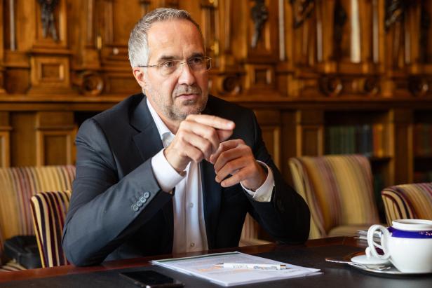 Ein Mann im Anzug sitzt an einem Tisch mit Unterlagen, Tasse und Smartphone und gestikuliert beim Sprechen.