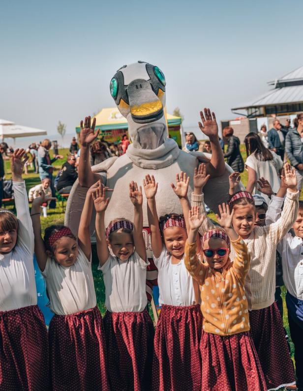 Mehrere Kinder in traditionellen Röcken und ein großes Vogelmaskottchen heben gemeinsam die Arme bei einer Veranstaltung im Freien.