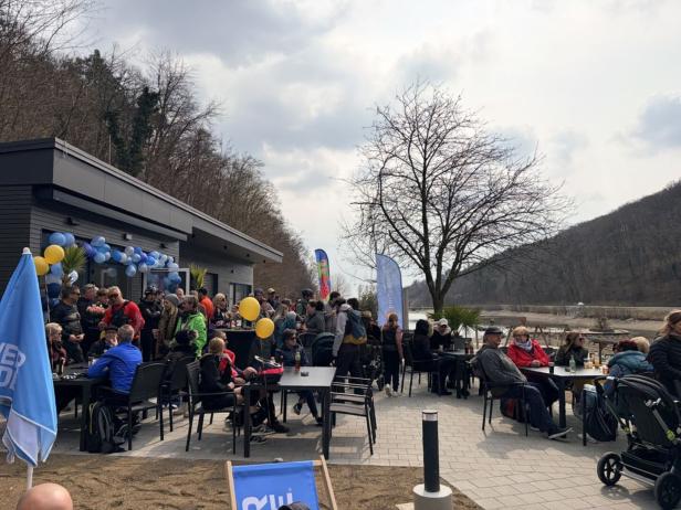 Viele Menschen sitzen und stehen an Tischen im Freien vor einem modernen Gebäude mit Luftballons, umgeben von Bäumen und Flusslandschaft.