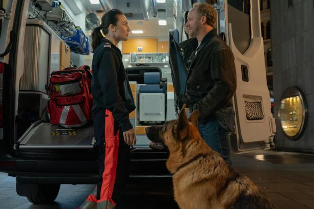 Eine Rettungssanitäterin spricht mit einem Mann neben einem Rettungswagen, während ein Schäferhund daneben sitzt.